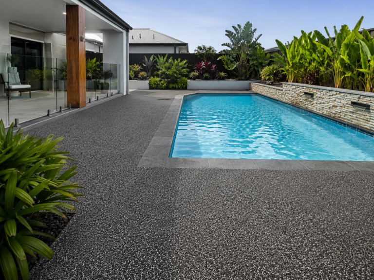 Freshly finished resin pool surround in Albany WA, showing a dark textured surface around a backyard swimming pool beside glass fencing, a covered patio, tropical garden beds and modern outdoor landscaping.