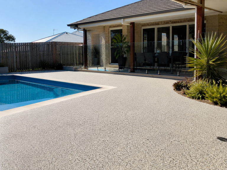 Freshly finished resin pool surround in Kwinana WA, showing a clean textured surface around a backyard swimming pool beside a modern patio, glass pool fencing, outdoor dining area and landscaped garden.