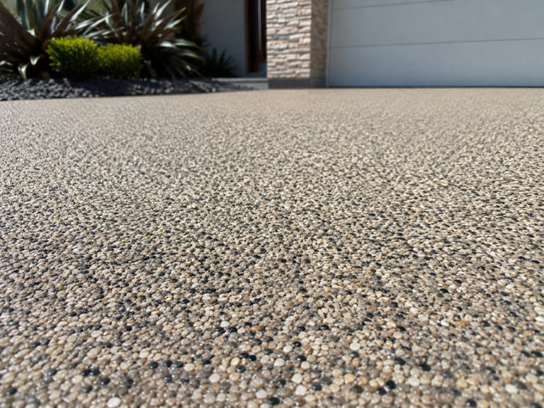 Close-up of a freshly finished resin driveway surface in Canning Vale WA, showing a textured pebble finish beside a modern home entry, garage door, stone exterior feature wall and landscaped garden beds.
